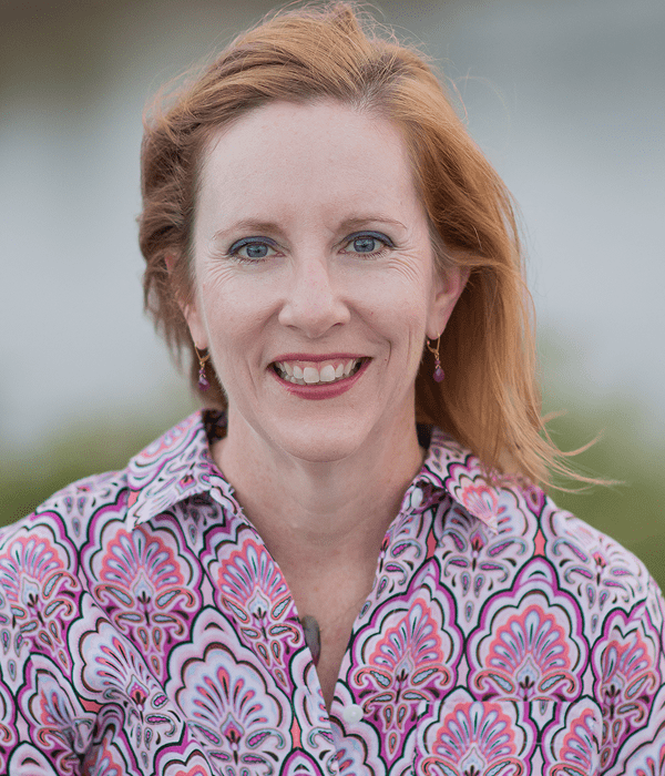 Smiling woman with red hair in a patterned shirt outdoors.